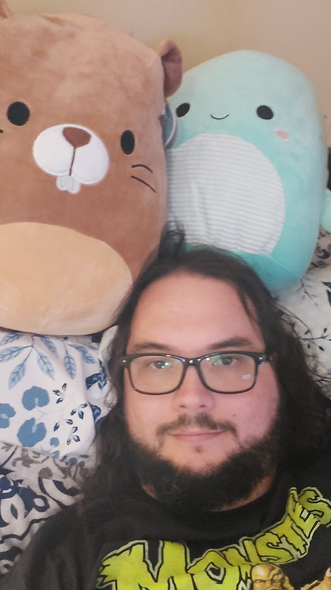 A man with long dark hair and a beard lies in bed with two Squishmallows, a beaver and a Nessie, photo 4